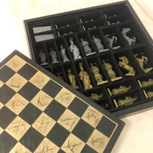 May include: A black and gold chessboard with a set of chess pieces in a black box. The chess pieces are made of a green material and are arranged in rows in the box.