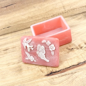 May include: A pink soap dish and soap bar set. The soap bar features white floral and butterfly patterns. The rectangular soap dish and soap bar are pink. The set is displayed on a wooden surface.