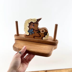 May include: Wooden unicorn-themed shelf with a light brown finish. The unicorn is painted with a cream-colored mane, a blue bow, and a floral design. Two wooden dowels are attached to the shelf.