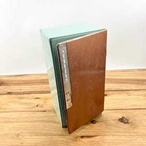 May include: A vintage metal file cabinet with a light blue exterior and a brown file folder. The file cabinet is slightly open, revealing the interior. The cabinet is on a wooden surface.