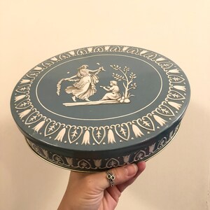 May include: A round, blue tin box with a white floral design. The lid features a scene of two women in white dresses with a floral background.