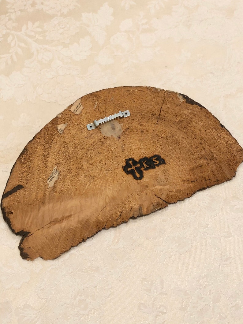 May include: The back of a wooden round sign with a metal hanger attached. The sign has a wood grain pattern and the words "PRS" are burned into the wood.