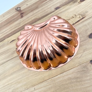 May include: A copper-colored seashell-shaped baking mold. The mold has a detailed, ridged design and a shiny finish. The kitchenware is made of metal and is designed for baking cakes or desserts.