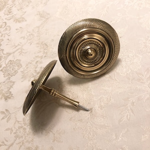May include: Two gold-toned metal curtain tiebacks with a decorative, circular design. The tiebacks have a screw-on attachment.