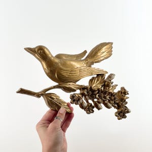 May include: A gold-colored bird sculpture perched on a branch with flowers. The bird is detailed with feathers and a beak, and the branch features leaves and blossoms. The sculpture is likely made of metal and is a decorative wall hanging.