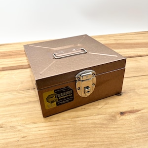 May include: A rectangular, metallic brown file box with a silver handle and clasp. The box has a label that reads "FILE AWAY CHECK FILE". The box is sitting on a wooden surface.