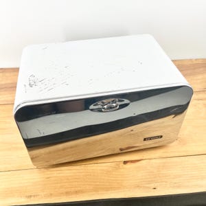 May include: A rectangular bread box featuring a white metal top with visible surface scratches, a polished chrome front panel with a central handle, and a light-toned wooden base. A small black label with the white text bread is on the front.