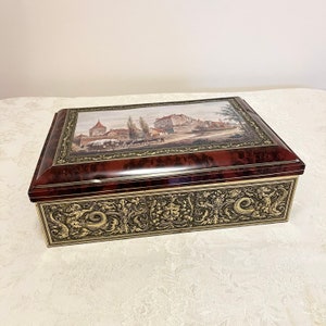 May include: A vintage, rectangular tin box with a red and gold finish. The lid features a painted scene of a medieval town with buildings and people. The box has an ornate, gold-colored design around the edges.