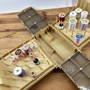 May include: An open, vintage sewing box with multiple compartments and a variety of spools of thread in various colors. The box is beige and has a clear lid with pins and buttons. The spools of thread are in shades of pink, white, and purple.