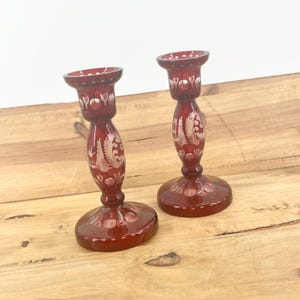 May include: Two ruby red glass candle holders. Each has a round base, a slender stem with etched floral designs, and a small cup at the top. The candle holders are displayed on a wooden surface.