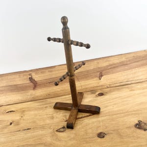 May include: A brown wooden mug tree with a central vertical post topped by a sphere, featuring two levels of horizontal arms with beaded detailing for hanging mugs, supported by a cross-shaped base made of four flat wooden pieces.
