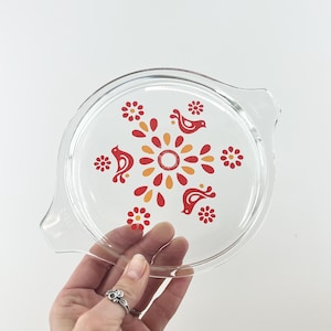 May include: Clear glass Pyrex lid with a decorative floral and bird design in red, orange, and yellow. The lid has a round shape with a handle on each side. A hand is holding the lid.