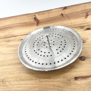 May include: A circular, stainless steel French press lid with a handle and numerous small holes. The lid is positioned on a light brown wooden surface.