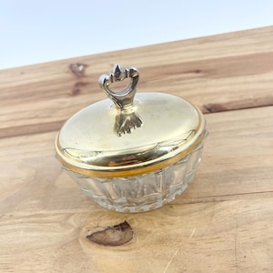 Peut inclure: Un petit pot à bijoux vintage en verre et doré, avec un couvercle décoratif. Le couvercle a une poignée argentée et un bord doré. La base en verre transparent a un design à facettes. Le pot est posé sur une surface en bois.
