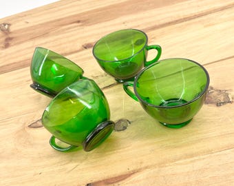 May include: Four green glass teacups with handles. The cups are a vibrant green color and have a rounded shape. Each cup sits on a small, dark base. The cups are arranged on a light brown wooden surface.