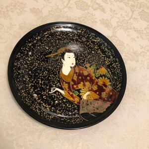 May include: A black ceramic plate with a gold and red geisha design. The geisha is wearing a traditional kimono and has a gold background.