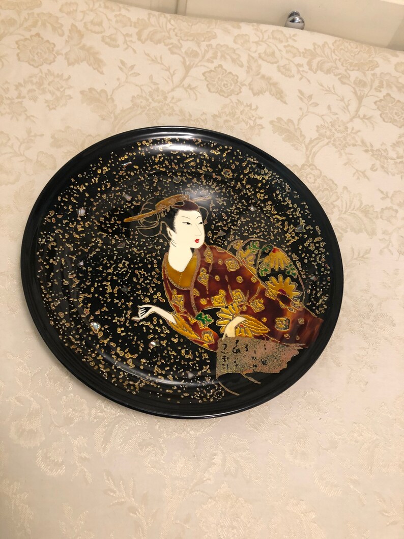 May include: A black ceramic plate with a gold and red geisha design. The geisha is wearing a traditional kimono and has a gold background.