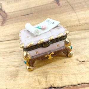 May include: A small, ornate trinket box with a hinged lid. The box is primarily pink with gold accents and a decorative scroll with the words "I love you" and a small pink rose. The box has a detailed, vintage design.