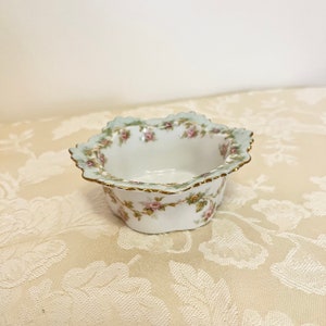 May include: A small, white porcelain bowl with a scalloped edge and a floral design. The bowl is decorated with pink and green roses and leaves. The bowl has a gold trim around the edge.