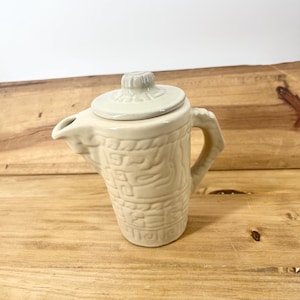 May include: A cream-colored ceramic creamer or small pitcher featuring a raised, textured geometric pattern around the body. It includes a matching removable lid with a small knob handle, a side handle, and a short pouring spout.