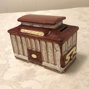 May include: A brown and white ceramic trolley piggy bank with a slot on the roof. The trolley has a sign that reads "Trolley" and the number "52" on the side.