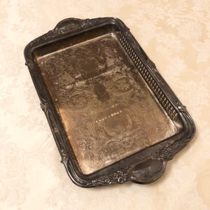 May include: A silver serving tray with an ornate design and two handles. The tray has a textured surface and a raised edge.