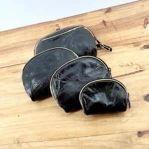 May include: Four black cosmetic bags of varying sizes are arranged on a wooden surface. Each bag has a rounded shape, a gold-colored zipper, and a textured surface. The bags are made of a shiny, possibly leather-like material.