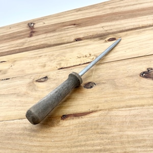 May include: A vintage steel and wood honing steel, used for sharpening knives. The handle is made of weathered gray wood, and the steel rod is silver. The tool is lying on a light brown wooden surface.