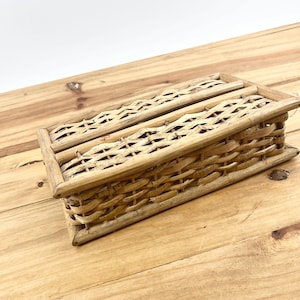 May include: A rectangular woven basket with a lid. The basket is made of light brown wicker and has a natural, rustic appearance. The weave pattern is visible, and the basket appears to be empty. The basket is sitting on a wooden surface.
