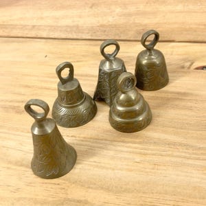 May include: Five antique brass bells with loop handles, each with a unique shape and etched floral patterns. The bells vary in size and design, showcasing a vintage aesthetic. The bells are arranged on a wooden surface.