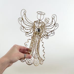 May include: A gold-colored wire angel Christmas tree topper with clear crystal accents. The angel has a halo, outstretched wings, and a central gem. The design features intricate wire detailing and decorative curls.