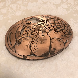 May include: A copper-colored metal trivet with a cutout design of fruit, including a pineapple, apples, and grapes. The trivet is oval-shaped and has a rustic, vintage look.