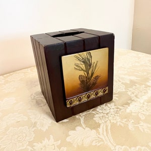 May include: A dark brown wooden tissue box cover with a gold and brown pine cone design. The design features a pine branch with pine cones and a decorative border.