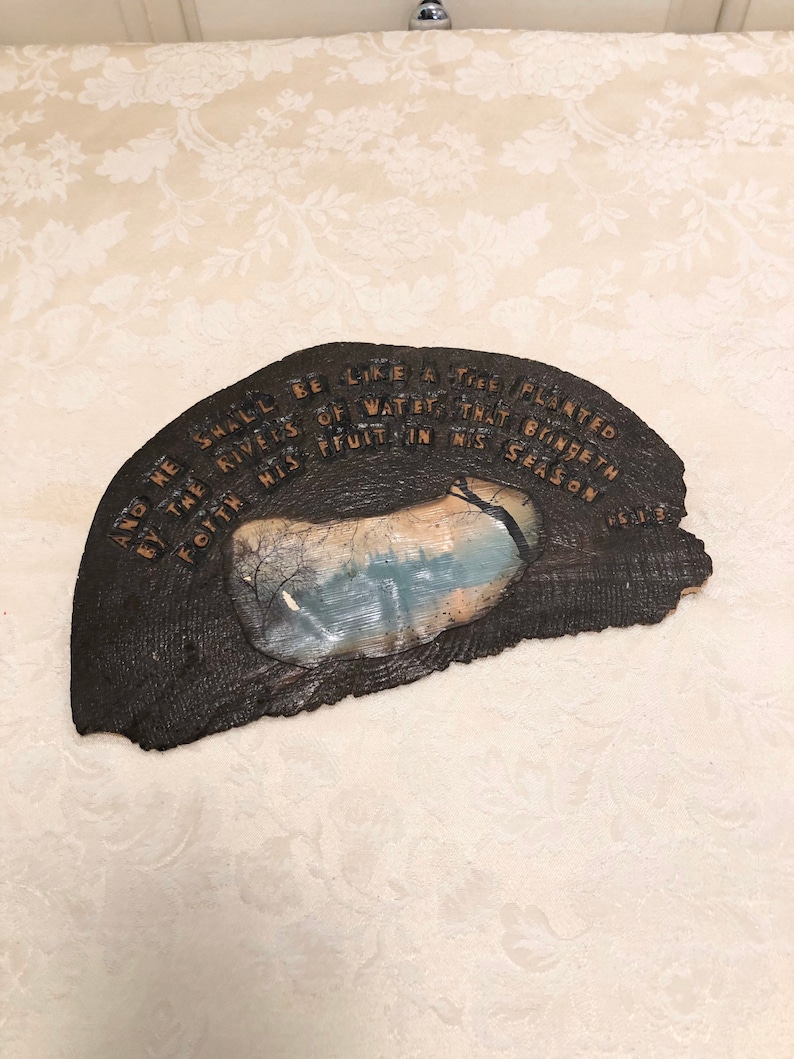 May include: A wooden plaque with a rustic brown finish and a painted scene of a lake with trees. The plaque has a quote from the Bible, "And he shall be like a tree planted by the rivers of water, that bringeth forth his fruit in his season."