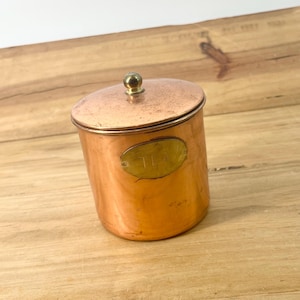 May include: A cylindrical copper tea canister with a matching lid and a small brass knob. The canister features an oval brass plaque on the front with the word TEA engraved in capital letters. The metal surface shows a warm, metallic finish.