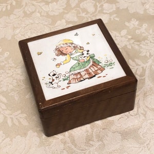 May include: A wooden box with a hinged lid. The lid features a colorful illustration of a girl in a green dress and a yellow hat playing with two dogs. The background is white with yellow butterflies.