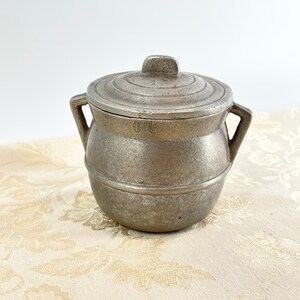 May include: A small, silver, metal pot with a lid. The pot has two handles and a slightly rounded shape. The pot is sitting on a white, floral patterned surface.