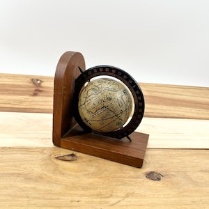 May include: A vintage-style globe bookend made of dark wood. The globe features a map design with the word "AFRICA" visible. A black metal ring with degree markings surrounds the globe. The bookend is on a wooden surface.