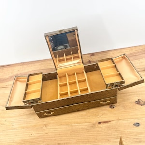 May include: A textured gold-tone jewelry box with an open lid featuring an interior mirror, multiple tiered compartments, and a bottom drawer. The interior is lined with a soft tan fabric, and the exterior has a subtle mottled pattern.