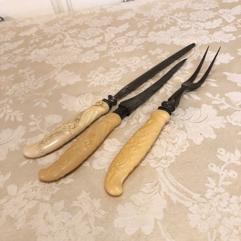 May include: Three antique carving tools with ivory handles. The tools are a carving knife, a smaller carving knife, and a carving fork.