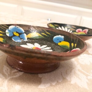 May include: Two small, hand-painted ceramic dishes with a black base and colourful floral designs. The dishes have a raised lip and a short, round foot.