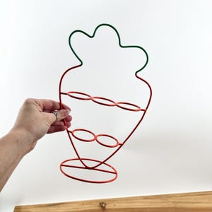 May include: A red and green wire egg holder shaped like a carrot. The holder has two tiers with three circles on each tier.
