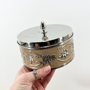 May include: A round, beige metal trinket box with a silver lid and floral design. The lid has a small, silver knob.