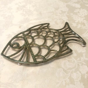 May include: A gold-toned metal fish-shaped hot pad with a decorative design. The hot pad has white rubber feet on the bottom.