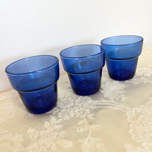 May include: Three cobalt blue glass cups with a fluted design. The cups are arranged in a row on a white tablecloth with a floral pattern.