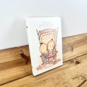 May include: A white photo album with a printed illustration of two children sitting in a large wooden rocking chair. The text "Betsy Clark PHOTO ALBUM" is printed in black at the top. The children are wearing blue and pink clothing.