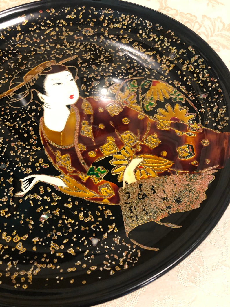 May include: A black lacquer plate with a gold and silver design of a woman in a kimono. The woman is surrounded by gold and silver dots and flowers.