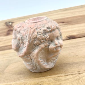 May include: A terracotta candle holder shaped like a cherub's head with a flower in its hair.
