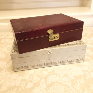 May include: Two vintage jewelry boxes stacked on top of each other. The top box is red with gold accents and a lock. The bottom box is white with a gold trim and a decorative pattern.