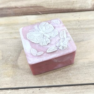May include: A small pink square box with a carved butterfly and floral design on the lid. The box is made of soapstone.